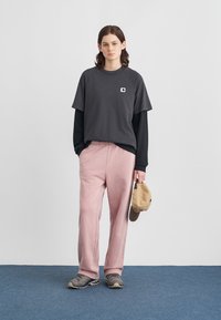 Graues Kurzarmhemd mit Logo, über einem schwarzen Langarmshirt getragen. Helle, rosaweite Hose, graue Turnschuhe und eine beige Kappe in der Hand.