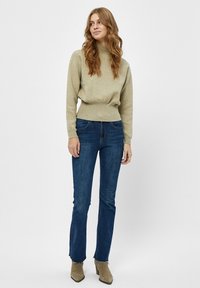 Pulover v svetlo olivno zeleni barvi z teksturiranim zaključkom, v kombinaciji s temno modrimi kavbojkami. Model nosi bež gležnjarje; nevtralen ozadje.