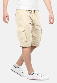Beige cargo shorts made of cotton, featuring multiple pockets and a belt with a dark buckle. Pairing with white mesh sneakers.