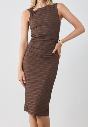 Femme portant une robe midi marron texturée sans manches avec un bracelet doré, debout devant un fond clair uni.