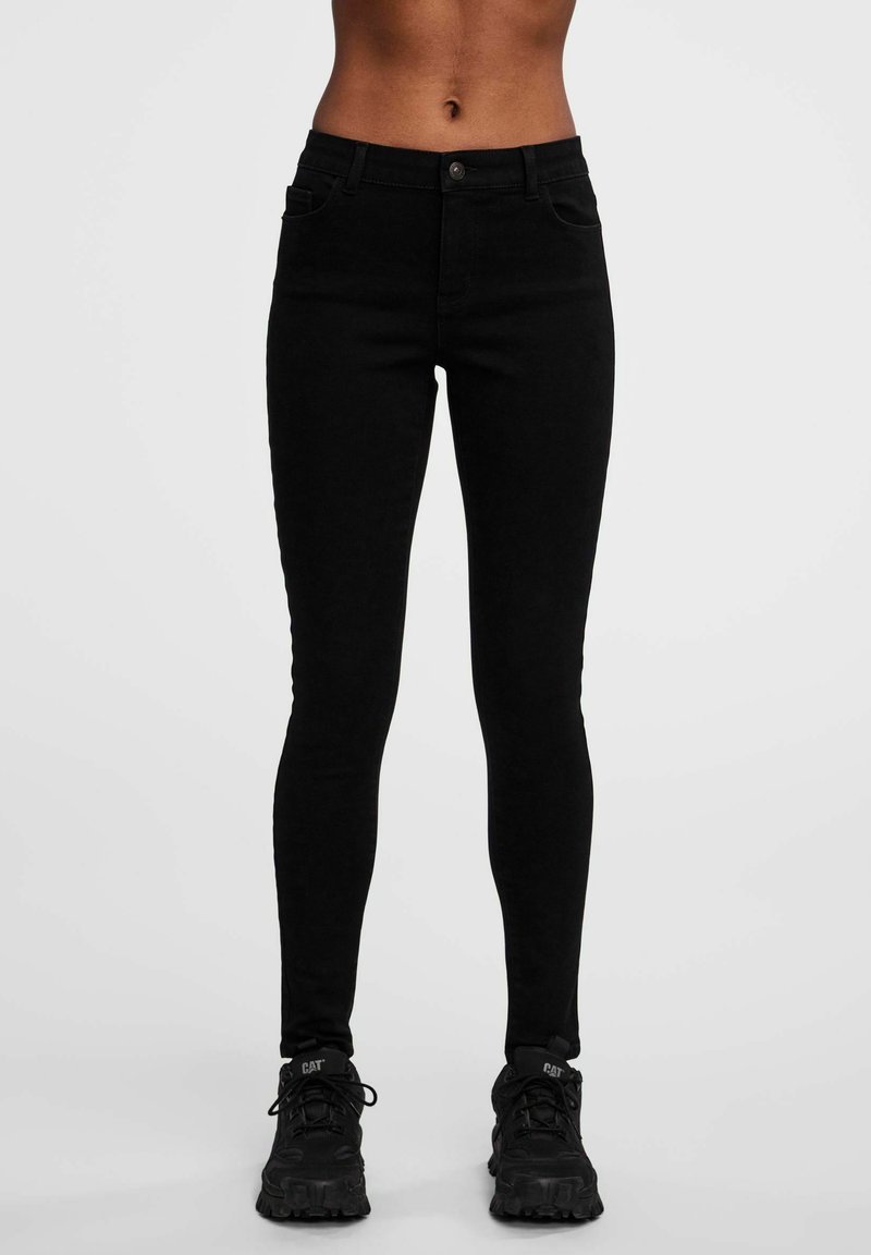 Jeans skinny noirs avec une taille haute, en denim. Comprend deux poches avant et une fermeture à bouton en métal. Portés avec des baskets noires.