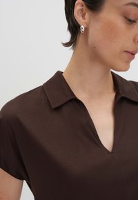 Chemise marron à col, avec un design en V, fabriquée en tissu lisse, dotée de manches courtes et de détails de couture subtils. Boucle d'oreille en argent visible.