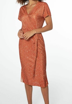 Kobieta w pomarańczowo-rdzawym sukience kopertowej z krótkimi rękawami i białym kropkowanym wzorem, z rękami splecionymi na wysokości talii na jednolitym tle.