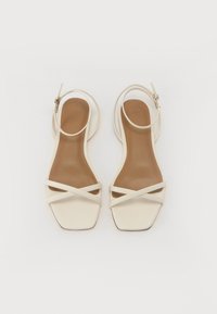 White strappy sandals feature a square toe design, flat heel, and crossed straps. The interior has a smooth, tan surface.