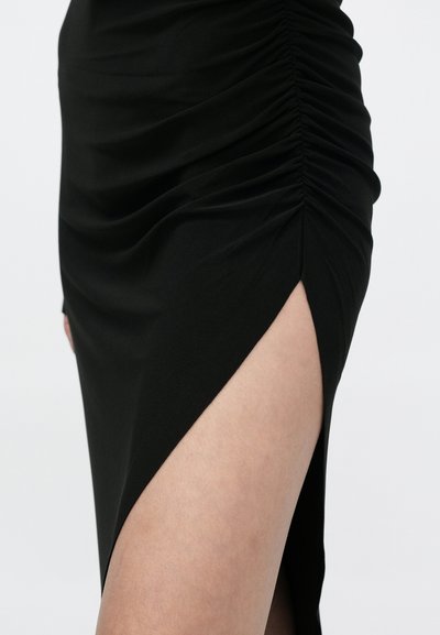 Μαύρη φούστα με ρυτιδωτό ύφασμα και υψηλή σχισμή στο μηρό που αποκαλύπτει γυμνό δέρμα, εμφανιζόμενη από το μέσο του μηρού έως τη μέση πάνω σε ένα απλό φωτεινό φόντο.