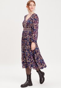 Vestido midi floral púrpura con mangas largas, escote en v y cintura elástica. Combinado con botas de tobillo negras que muestran patrones intrincados.