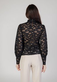 Schwarze Spitzenbluse mit langen Puffärmeln, hohem Kragen, welligem Saum und Knopfverschluss hinten. Verfügt über ein florales Muster und einen subtilen Glanz.