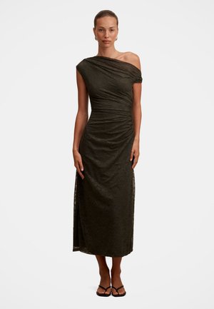 Donna che indossa un vestito midi scuro, monospalla, aderente con una sottile texture e sandali neri aperti in punta, in piedi su sfondo bianco.