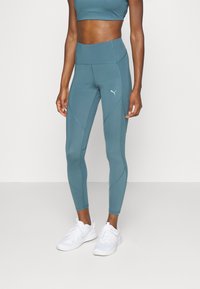 Leggings deportivos color teal con cintura alta, que presentan costuras triangulares y un pequeño logo; combinados con zapatillas de correr blancas.