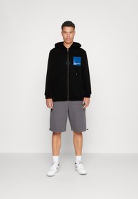 Crna zip-up hoodica s plavim kvadratnim logotipom na džepu, uparena sa sivim šortsevima i bijelim tenisicama. Glatka tekstura, ležeran dizajn.