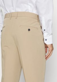 Beige színű nadrág sima textúrával, hátul fekete gombos zsebekkel és slim fit szabással, fehér inggel párosítva.