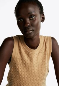 Beige gebreide mouwloze top met een kabelpatroon, ronde halslijn en geribbelde randen, met een gladde textuur en een aansluitend ontwerp.