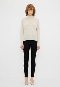 Lichtbeige trui met een hoge hals en een textuur van stippenpatroon, gecombineerd met zwarte leggings en blushkleurige hakken. Eenvoudig ontwerp, getailleerde vorm.