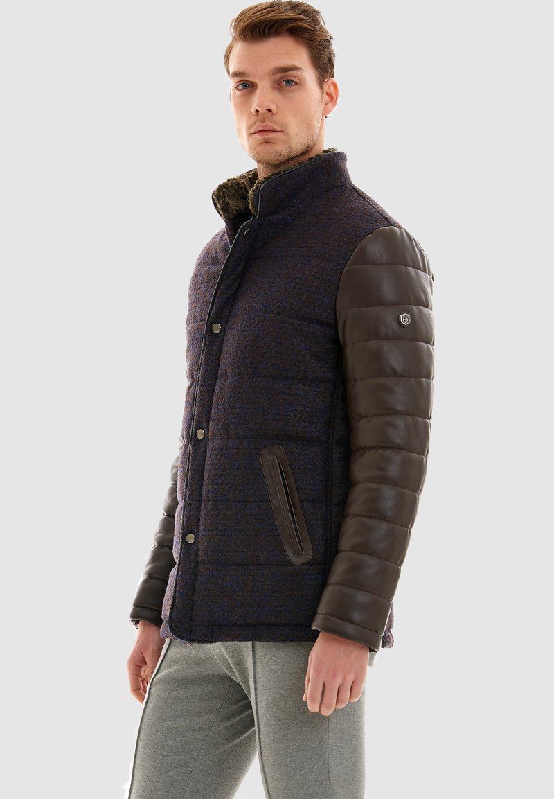 Vadderad jacka med en texturerad marinblå kropp och bruna lädersleeves, med en ståkrage och två sidofickor.