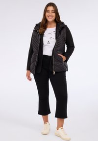 Černá prošívaná vesta s kapucí, se zipem a bočními kapsami, vrstvená přes bílé tričko s krátkým rukávem. Nosené s černými kalhotami a krémovými teniskami.