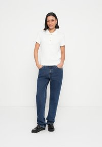 Polo blanc en coton avec un col et trois boutons dorés, associé à un jean en denim bleu ample et des chaussures noires.
