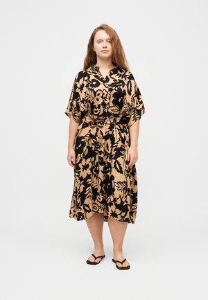 Γυναίκα με μακριά κοκκινωπά μαλλιά που φοράει μπεζ midi φόρεμα με μεγάλα μαύρα ανθισμένα σχέδια και μαύρα σαγιονάρες, στέκεται μπροστά σε απλό φόντο.