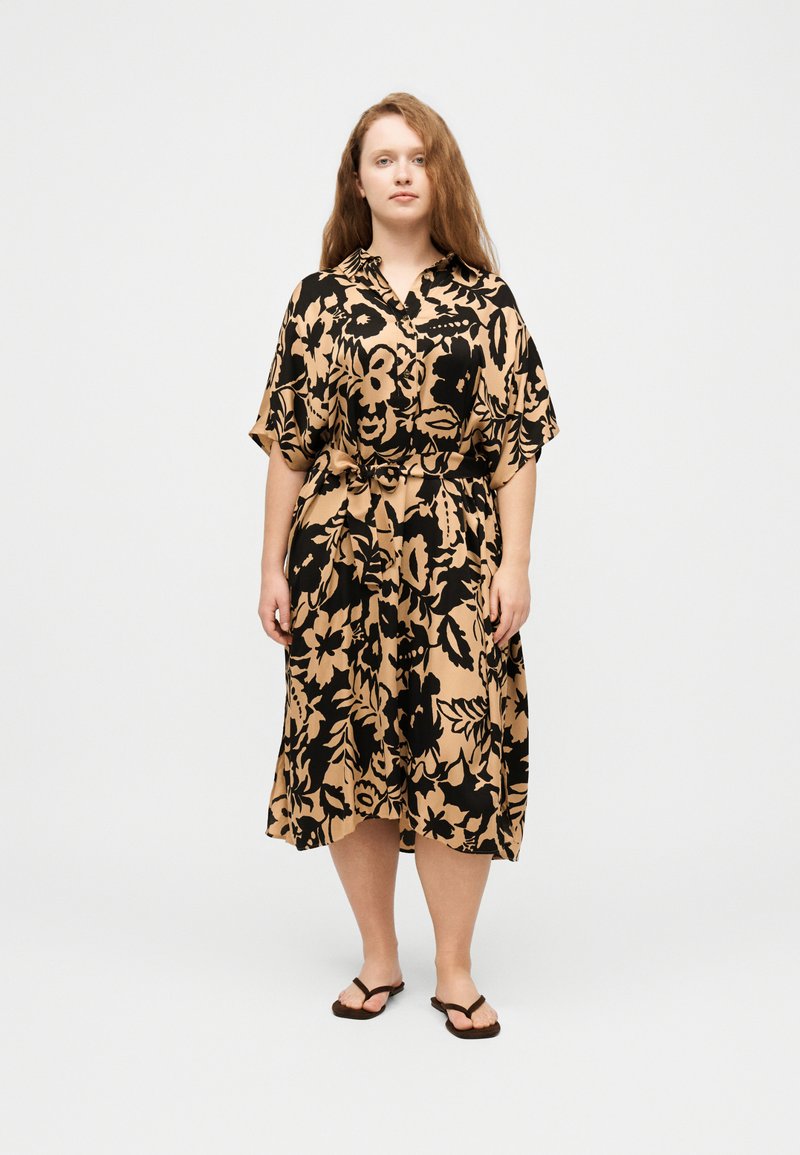 Γυναίκα με μακριά κοκκινωπά μαλλιά που φοράει μπεζ midi φόρεμα με μεγάλα μαύρα ανθισμένα σχέδια και μαύρα σαγιονάρες, στέκεται μπροστά σε απλό φόντο.