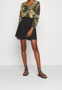 Top lungo a maniche con fiori verdi e scollo a V, abbinato a una gonna nera svasata e sandali neri con borchie. Design semplice e aderente.