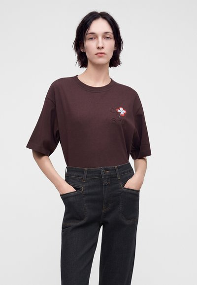 Γυναίκα με κοντά σκούρα μαλλιά φοράει καφέ oversized t-shirt με μικρή σχεδίαση λουλουδιού και μαύρο τζιν ψηλόμεσο με τα χέρια στις τσέπες.