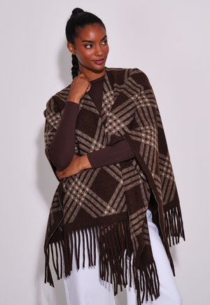 Vrouw met donker gevlochten haar draagt een bruine en beige geruite sjaal met franjes over een donkere top met lange mouwen en witte broek, staand tegen een lichte achtergrond.