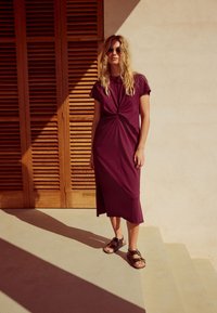 Robe trapèze grenat avec détail noué à l'avant et manches courtes. Associée à des sandales plates marron. Fond composé de lattes en bois.