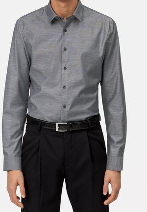 Homme portant une chemise grise cintrée à boutons, rentrée dans un pantalon noir plissé avec une ceinture noire.