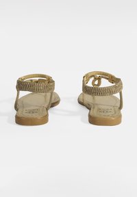 Goldene Flachsandalen mit einem geflochtenen Riemendesign, strukturierten Oberflächen und einer flachen braunen Sohle. Offene Ferse und Vorderseite für Belüftung.