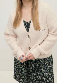 Žena v krémovém pleteném svetru s knoflíky přes černé květinové šaty, ukazující ruce se prsteny a s dlouhými rovnými vlasy.