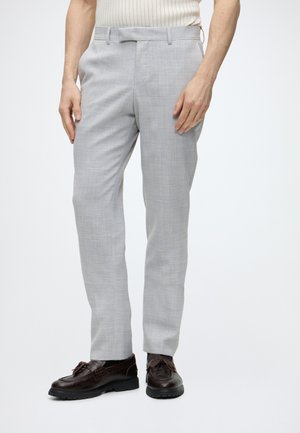 Homme portant un pantalon de costume gris clair, des mocassins en cuir marron foncé avec pampilles et un haut blanc côtelé sans manches, debout devant un fond blanc.