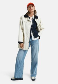 Weißer Außenmantel mit marineblauem Kragen, über einem marineblauen Cardigan und einem weißen Hemd getragen, kombiniert mit hellblauen weiten Jeans und schwarzen Schuhen.