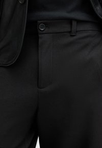 Pantalones negros a medida fabricados en una tela suave que presenta una textura sutil, un bolsillo frontal y un cierre de botón al lado de la cintura.