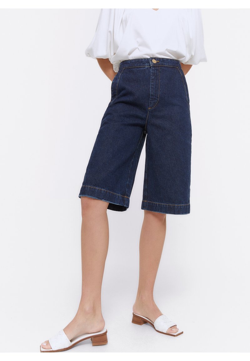 Henkilö, joka on pukeutunut korkeavyötäröisiin tummaan farkuisiin polvipituisiin housuihin ja valkoisiin avokärkisiin korkokenkiin, seisoo valkoista taustaa vasten.