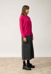 Fuchsia Pullover mit halbem Reißverschluss, kombiniert mit einem langen grauen Denim-Rock. Das Modell trägt schwarze Schnürschuhe und Socken. Schlichtes, sauberes Design.