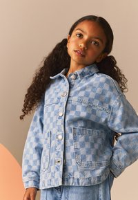 Veste en denim bleu clair avec un motif à damiers, dotée d'un col, de poches avant et de boutons en métal argenté. Portée de manière décontractée.