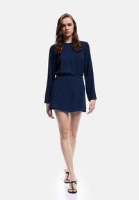 Marineblauwe jurk met lange mouwen, een strakke taille en een korte rok. Glad stof met minimale details, gecombineerd met zwarte sandaaltjes met banden.