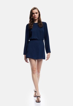 Marineblauwe jurk met lange mouwen, een strakke taille en een korte rok. Glad stof met minimale details, gecombineerd met zwarte sandaaltjes met banden.