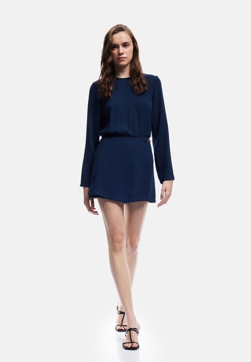 Marineblauwe jurk met lange mouwen, een strakke taille en een korte rok. Glad stof met minimale details, gecombineerd met zwarte sandaaltjes met banden.