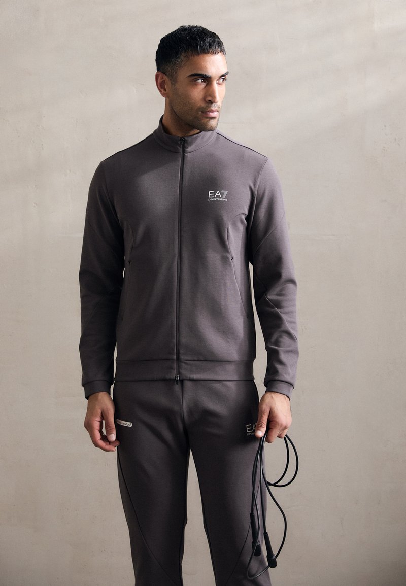 Tuta grigia in tessuto liscio, con giacca con zip, collo alto e logo, abbinata a pantaloni coordinati e una corda per saltare.