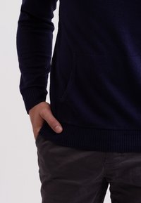 Mörkblå stickad tröja med ribbat nederkant och manschetter, med en framficka. Parat med mörkgrå byxor som visar en slät tygstruktur.