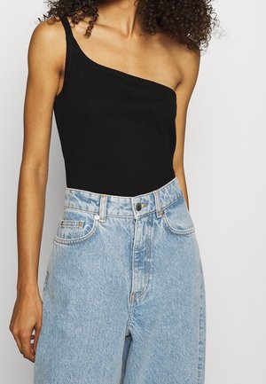 Frau trägt ein schwarzes ärmelloses Top mit einem Schulterriemen, kombiniert mit hoch taillierten hellblauen Jeans vor einfachem Hintergrund.