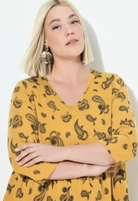 Donna bionda con taglio a caschetto indossa un vestito giallo con motivo paisley e orecchini a forma di foglia d'oro, in piedi con le braccia incrociate su uno sfondo bianco.