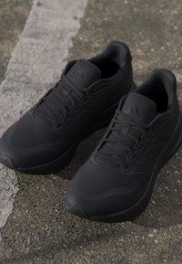 O pereche de pantofi de alergare Adidas negri cu șireturi, așezați pe asfalt texturat lângă o linie albă ștearsă.