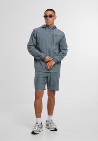 Lehká šedá bunda s kapucí a zipovou kapsou; doplněná o odpovídající šortky. Doplňky zahrnují sportovní sluneční brýle a síťové tenisky.