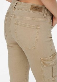 Pantalones cargo beige de corte Slim, con bolsillos laterales, bolsillos en la parte posterior y una etiqueta de marca de cuero marrón en la cintura.