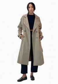 Manteau beige à double boutonnage, grand revers cranté et manches larges ; porté sur un crop top bleu marine et un pantalon foncé.