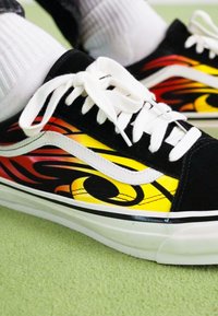 Zapatillas de lona negras con diseños de llamas amarillas y rojas, cordones blancos y una franja lateral blanca. Suela de goma texturizada.