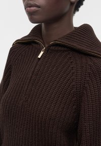 Gros plan d'une personne portant un pull en grosse maille marron foncé avec un grand col et une fermeture éclair dorée partiellement fermée.