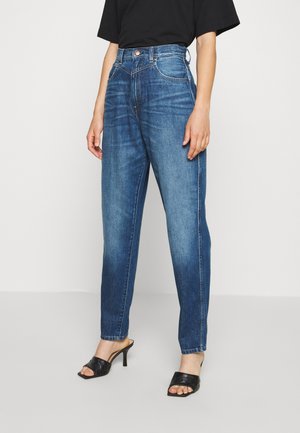 Kobieta ubrana w niebieskie jeansy z wysokim stanem, czarną koszulkę i czarne sandały na obcasie z odkrytymi palcami, stojąca na jednolitym tle.