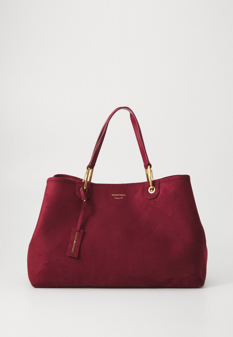 Burgundská semišová taška s zlatistým kovanim, dvojitými rúčkami a pripevnenou visačkou. Hladký povrch a priestranný dizajn pre všestranné využitie.